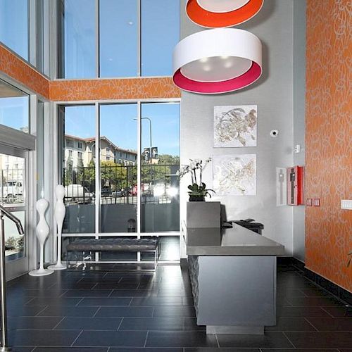 A lobby area with modern decor, featuring a reception desk, seating, bright lighting, orange wallpaper, and large windows providing natural light.