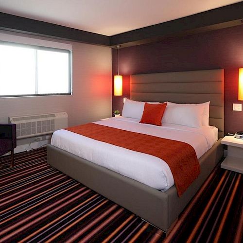 The image shows a modern hotel room with a large bed, striped carpet, purple accent wall, bedside tables, and warm lighting from two hanging lamps.