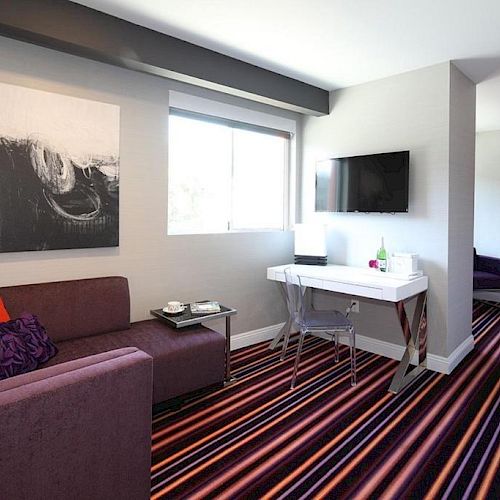 A modern living room features a purple couch, striped carpet, abstract artwork, a flat-screen TV, and a small desk near a window.