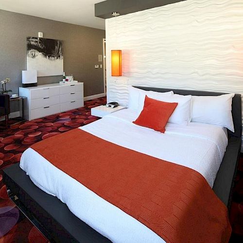 A modern hotel room with a double bed featuring white and orange bedding, unique orange light fixtures, and contemporary furniture.