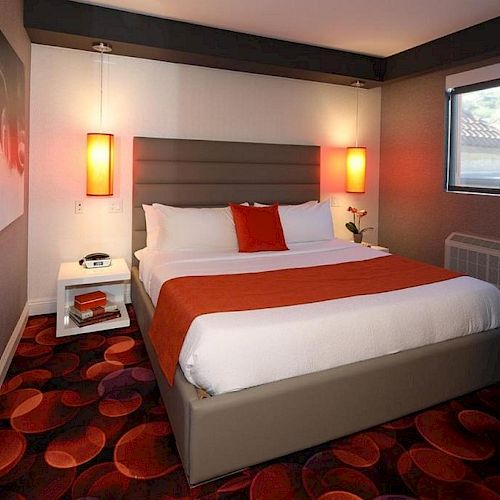 A modern hotel room with a double bed, two bedside tables with lamps, a window, a patterned carpet, and a mirror reflecting part of the room.