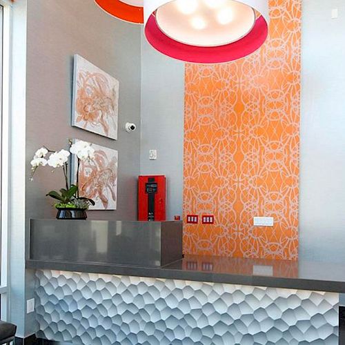 A modern reception area with orange patterned wallpaper, a white textured desk, wall art, a hanging lamp, and plants for decoration.