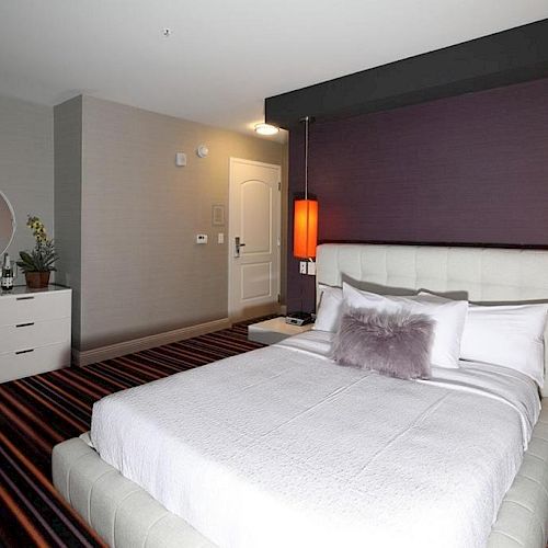 A modern bedroom featuring a double bed with white bedding, purple accent wall, red hanging lights, a dresser with a mirror, and striped carpet.