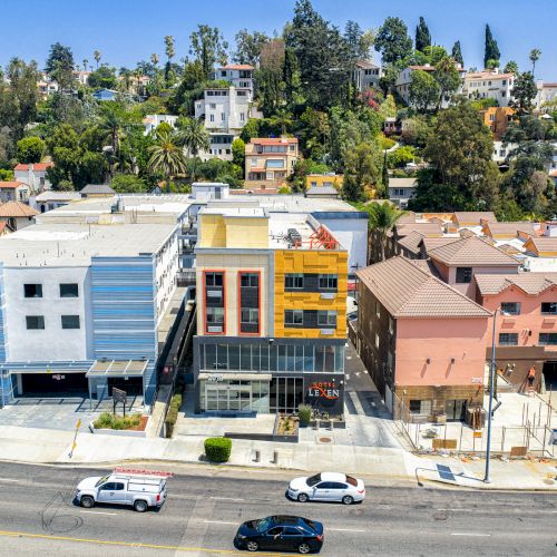 Aerial view of a street with colorful buildings, parked cars, and a hilly backdrop with trees and houses in the background.