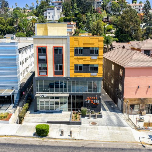 The image shows a row of modern buildings with different color schemes, set against a backdrop of trees and a slight hill.