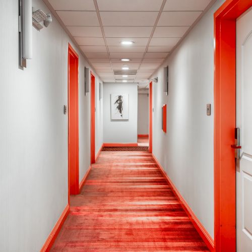 A well-lit hotel hallway with orange and white decor, several doors, and a painting at the end.