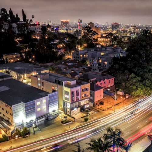 A cityscape at night with buildings, streets, trees, and blurred car lights.