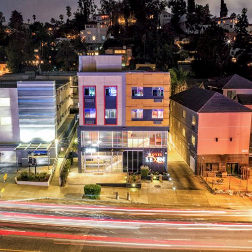 A nighttime urban scene with buildings, illuminated windows, and motion blur from car lights on the street, with a hillside backdrop.
