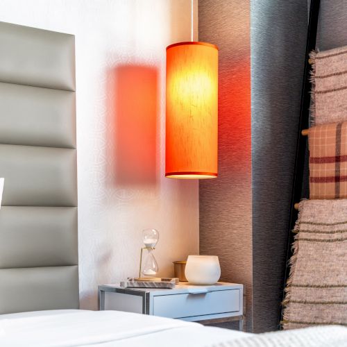 A cozy bedroom scene featuring a soft headboard, a warm orange lamp, a bedside table with decor, and neatly stacked blankets on a rack.