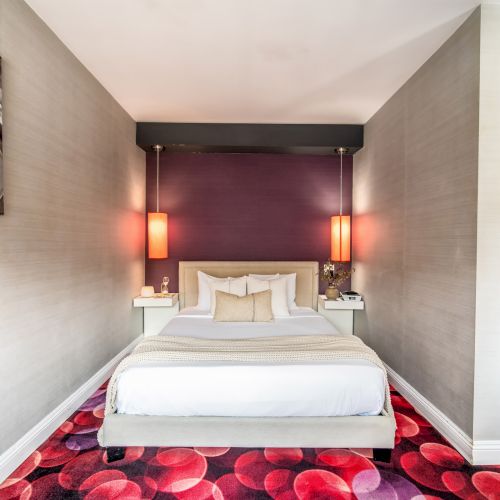 The image shows a modern bedroom with a bed, decorative pillows, vibrant red and pink patterned carpet, and wall lamps on either side.