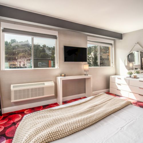 A modern bedroom with red carpet, bed, wall-mounted TV, dresser with mirror, windows, and a blanket ladder for decor.