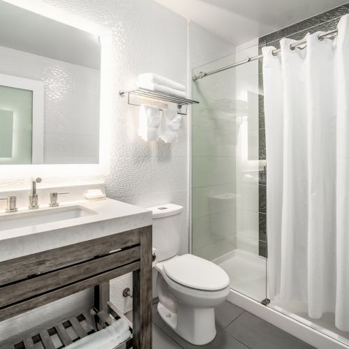 The image shows a modern bathroom with a sink, mirror, toilet, and shower with a white curtain. Towels are neatly arranged on a shelf.