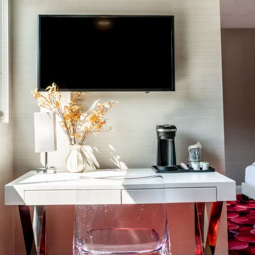 A modern room with a TV, table, vase of flowers, and coffee maker. There's a bed in the background and a window on the left.