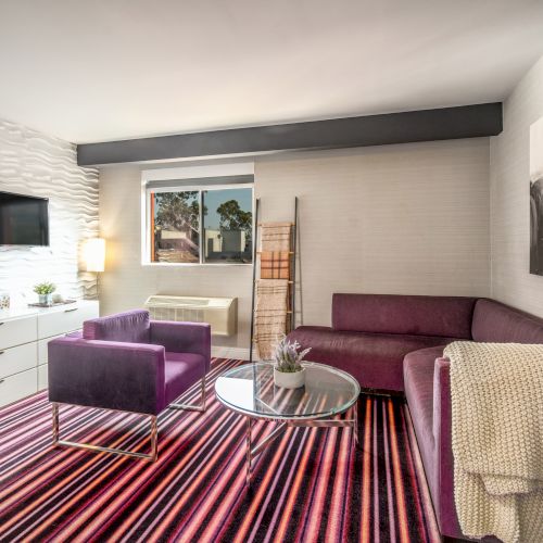 A modern living room with purple furniture, a striped rug, wall art, TV, and a window allowing natural light into the space.
