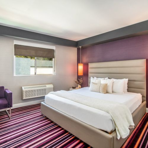 A modern bedroom with a striped carpet, purple walls, a large bed, an armchair, two pendant lights, and a window with a blind.