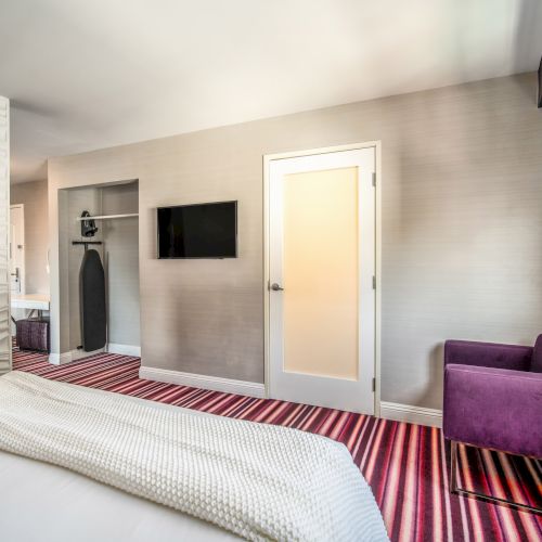 A modern hotel room with a striped carpet, purple chair, TV, and closet, featuring a minimalist design and neutral color palette.