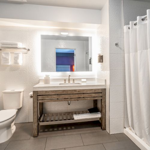 A modern bathroom with a toilet, wooden vanity, illuminated mirror, and a shower with a white curtain and towel rack.