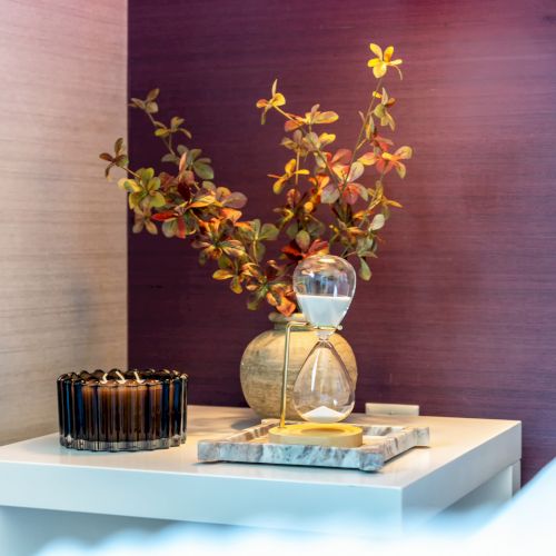 A bedside table with decorative items: a vase with flowers, a candle, and a book, set against a purple wall.