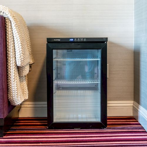 A small, black mini-fridge stands next to a sofa draped with a knitted blanket, set on a striped carpet against a light-colored wall.