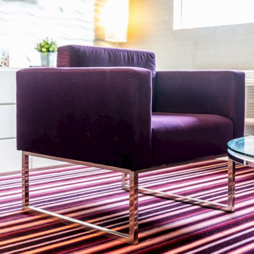 A modern room features a purple armchair on a striped carpet with a glass table nearby, illuminated by a window's natural light.