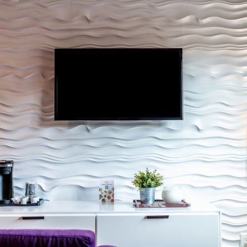 A modern room features a TV on a textured wall, with a cabinet holding a coffee maker and plants, and a floor lamp nearby.