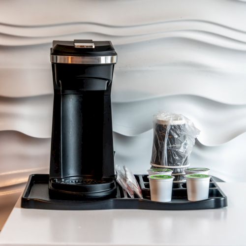 A single-serve coffee maker sits on a tray with cups and wrapped accessories against a wavy white textured wall.