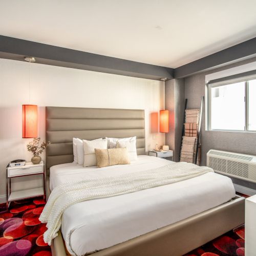 A modern bedroom with a large bed, colorful carpet, side tables, two pendant lights, and a window with blinds, creating a cozy atmosphere.