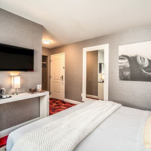 The image shows a modern hotel room with a bed, wall-mounted TV, art, and a desk with a lamp and coffee maker, decorated in neutral tones.